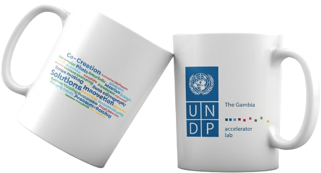 White ceramic mug printed with the UNDP The Gambia Accelerator Lab logo on one side and a multicolour word cloud on the other.